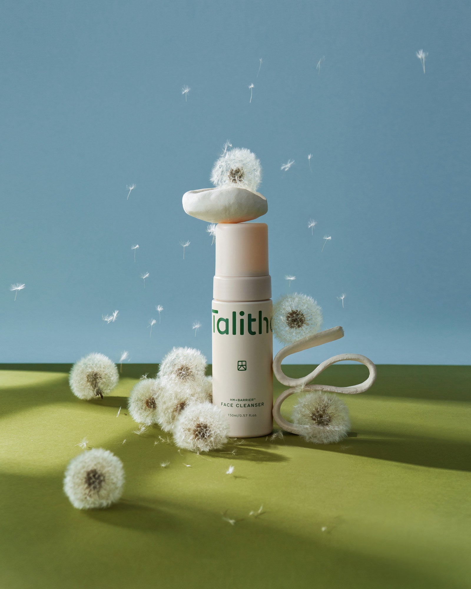Face Cleanser among white dandelion puffs on a green and blue set